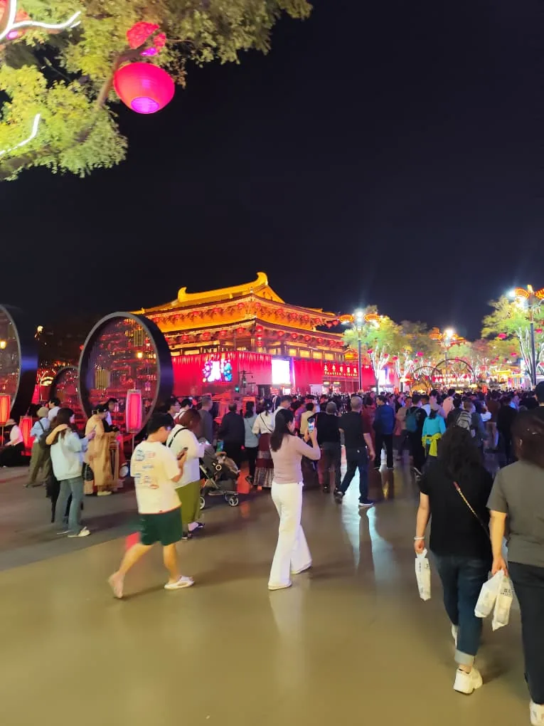 Plaza nocturna frente a un pabellón tradicional chino iluminado, con faroles y numerosos visitantes, evocando la muralla iluminada de Xi'an.