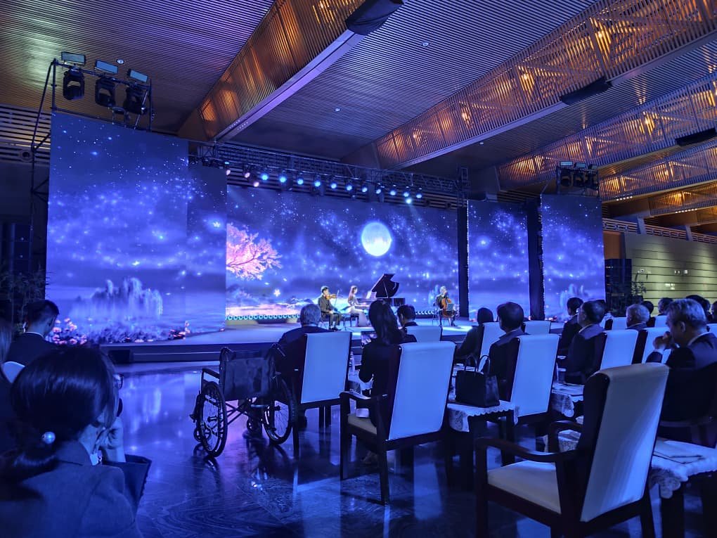 Gala cultural con músicos en escenario y una gran pantalla LED que proyecta un paisaje estrellado con luna y cerezo, ante un público en un salón moderno de China.
